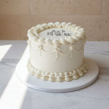 Elegant white buttercream celebration cake with piped detailing and gold lettering, displayed on a marble surface with soft natural light at Prestige Patisserie in North London.