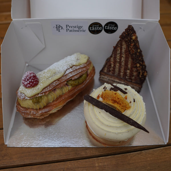 Three artisan desserts—layered Mountain Cake, Banoffee Tart topped with honeycomb shards, and a Pistachio & Raspberry Éclair—neatly arranged in a kraft-paper gift box.