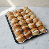 A tray of mini brioches filled with chive cream cheese ans smoked salmon, arranged on a white lace doily over a gray surface.