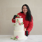 Geanina from Prestige Patisserie holding a three-tiered wedding Medovik-Honey Cake with floral decorations against a beige wall.