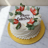A round white frosted cake on a gold cake board, decorated with sliced strawberries and green leaves around the top, with 'HAPPY CHRISTMAS' written in icing. The cake is placed on a white marble surface against a light beige wall.