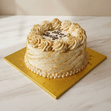 A round cake with beige and white frosting, piped rosettes around the top, and a gold board underneath, photographed on a light marble surface against a beige wall.
